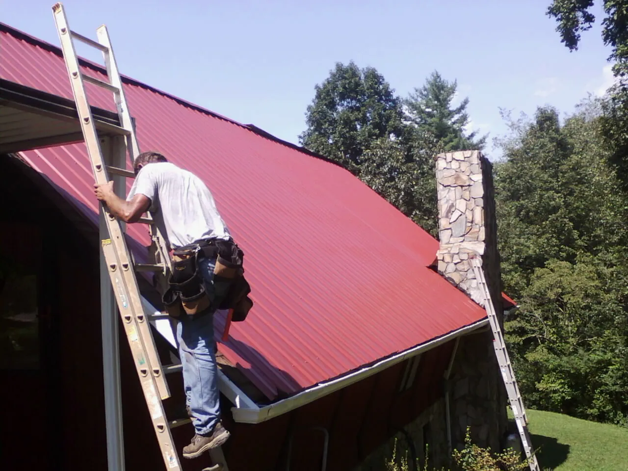Roofer on a ladder beside a red metal roof and stone chimney — Soffit & Fascia Repair by Roofing Brothers in Maury City